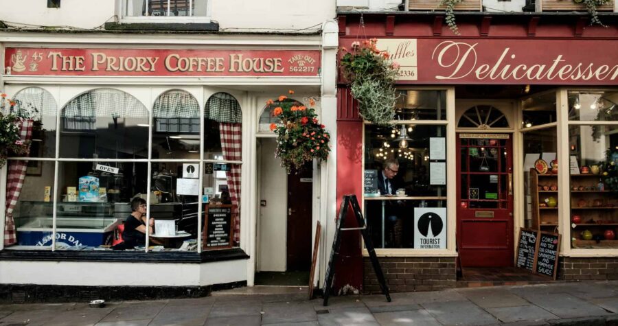 A vibrant street scene showing two shops, "The Priory Coffee House" on the left with a woman sitting by the window and "Delicatessen" on the right with a man enjoying a beverage, both shops have plants hanging above the entrances, a tourist information sign, and a clear view of the cobbled street outside