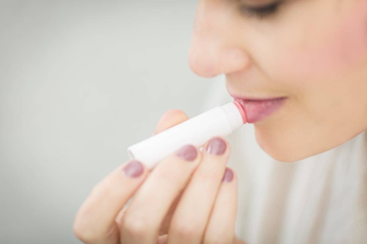 Woman applying pink lip balm, close-up, soft focus, white tube, natural makeup, smooth lips, gentle expression, light background