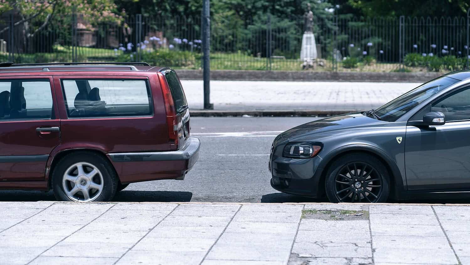 A red Volvo and a grey car parked side by side on a city street, the vehicles are in close proximity, with the focus on their contrasting appearances, the background includes a statue and green plants, indicating a well-maintained urban environment