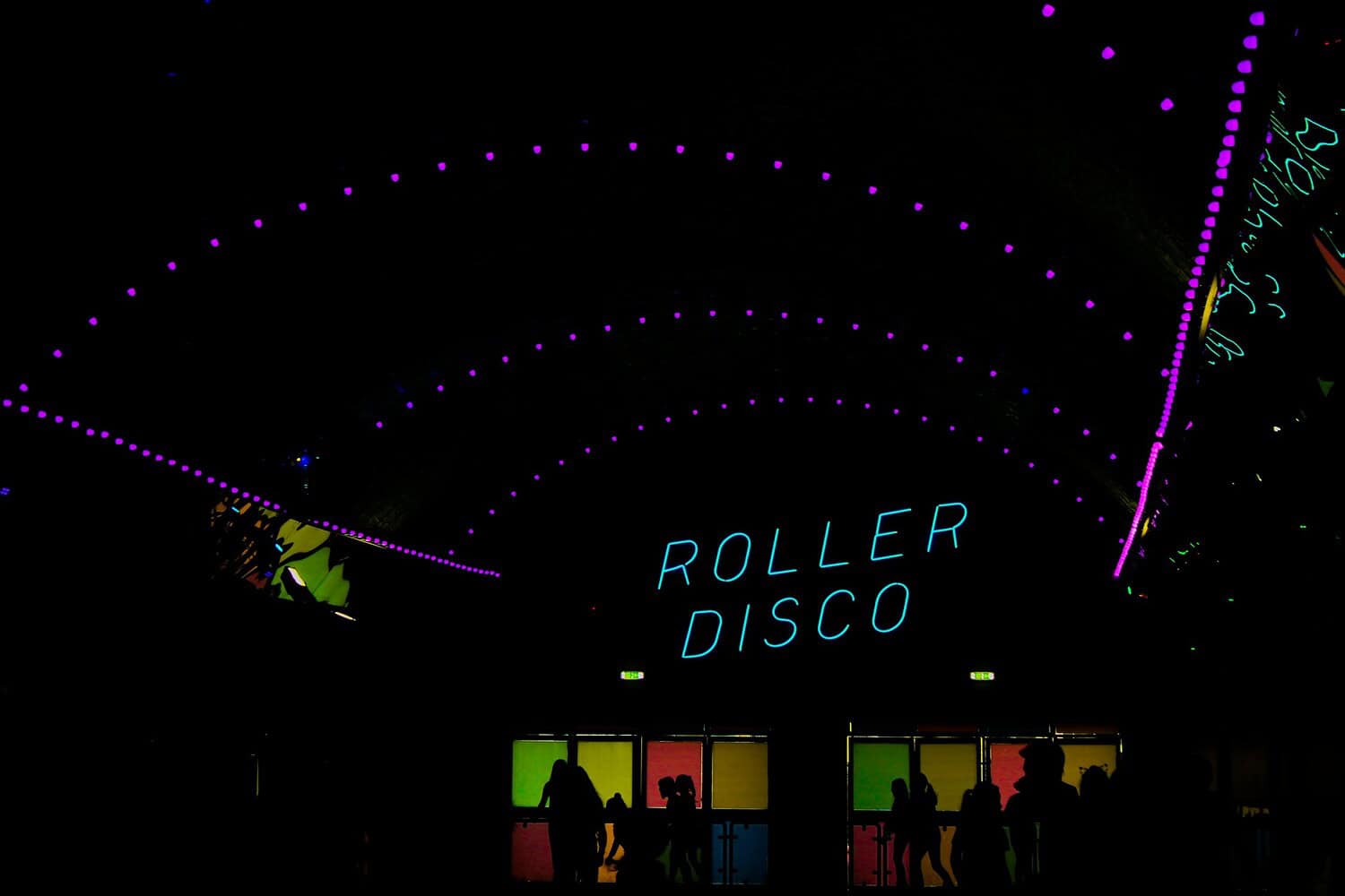 Roller disco sign glowing in neon blue, colorful lights illuminating the entrance, silhouettes of people moving near the door, bright purple and neon lights arching over the space, a vibrant, lively atmosphere in the roller disco