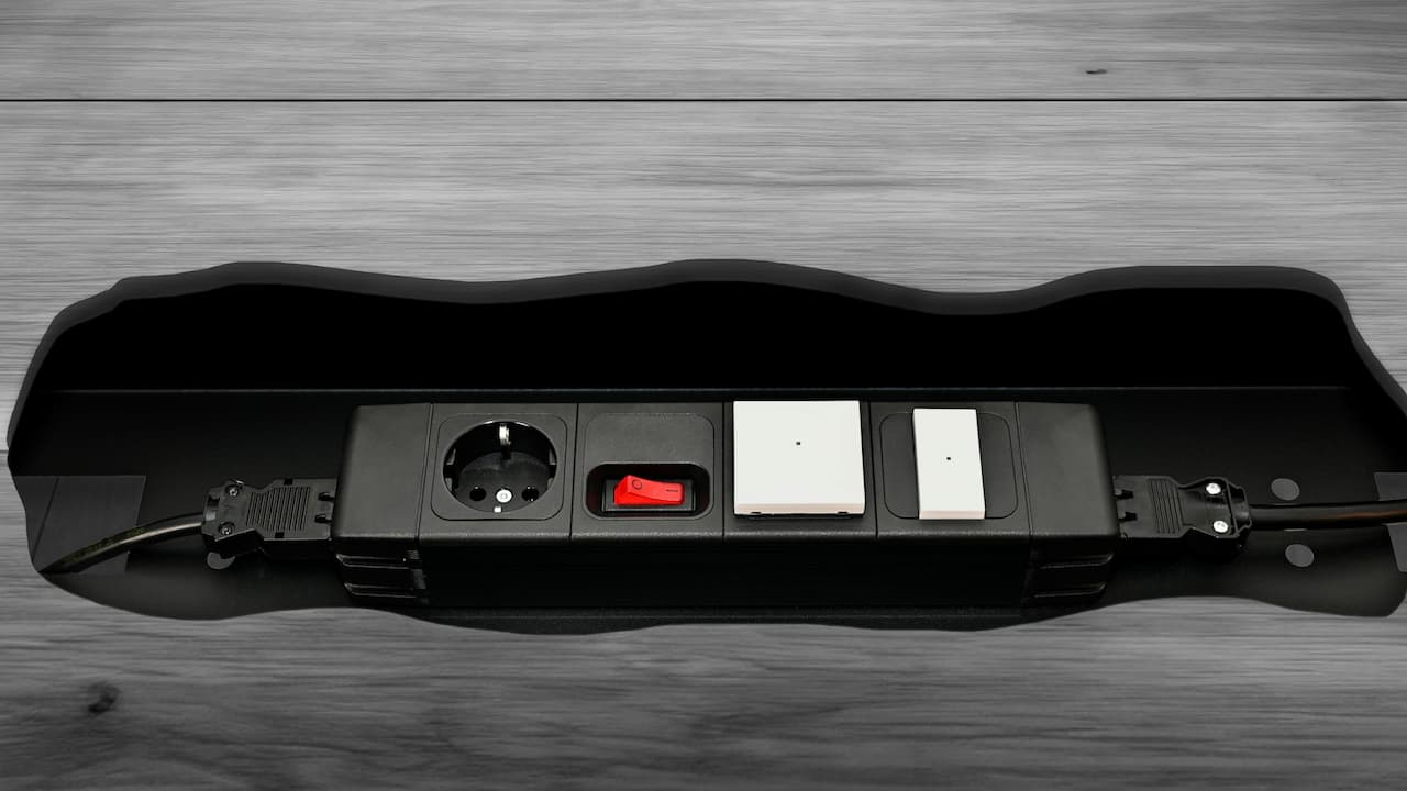 Black desk smart power strip with European electrical outlet, red power switch, two white modules, and cable management system on wooden surface