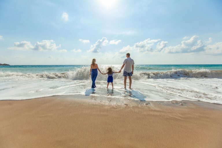 A family of three is holding hands, standing together on a beach near the ocean, the sun is shining, waves are gently washing up on the shore, the child is in the middle, and the parents are on each side, enjoying a sunny day, the soft sand and water are visible beneath their feet