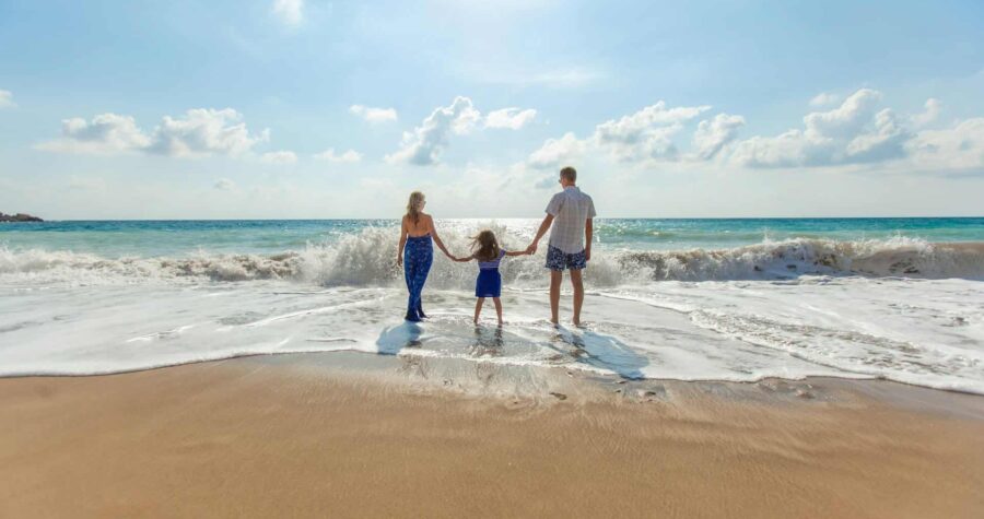 A family of three is holding hands, standing together on a beach near the ocean, the sun is shining, waves are gently washing up on the shore, the child is in the middle, and the parents are on each side, enjoying a sunny day, the soft sand and water are visible beneath their feet