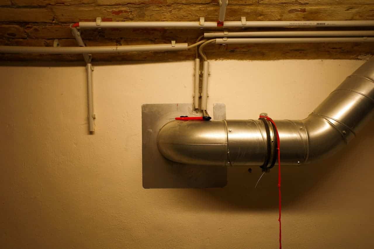 Metal insulation blower duct mounted on beige wall with white PVC pipes above, used for installing loose insulation