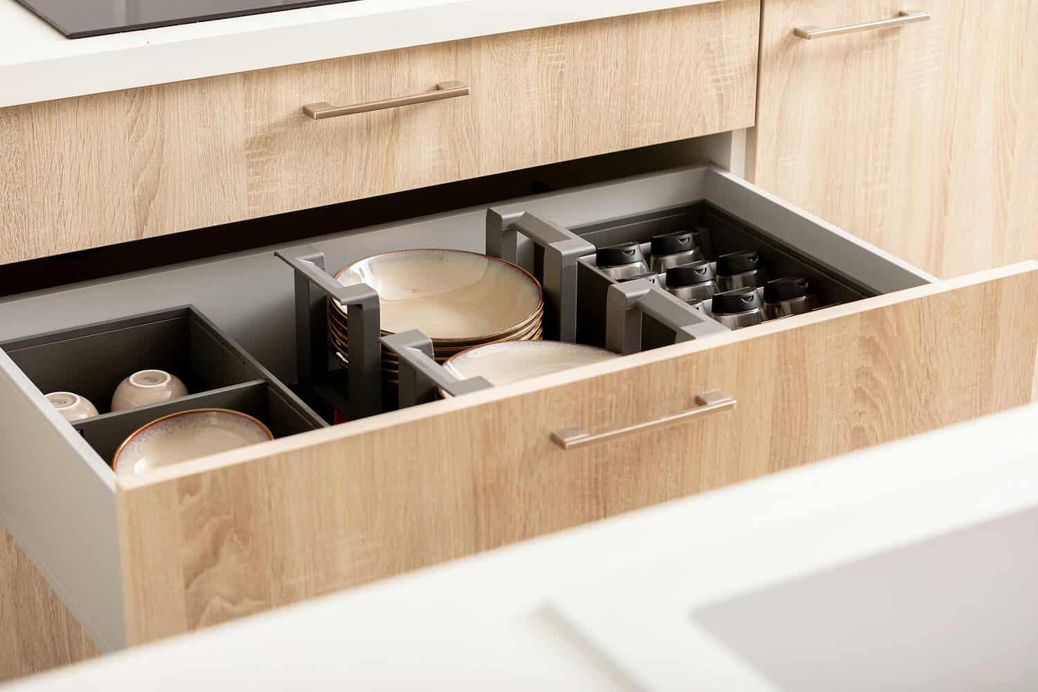 A neatly organized kitchen drawer, containing stacked plates secured in dividers, small bowls in separate compartments, and spaces for cups, clean and orderly with light-colored wood surroundings and modern drawer hardware, offering efficient storage for dishware