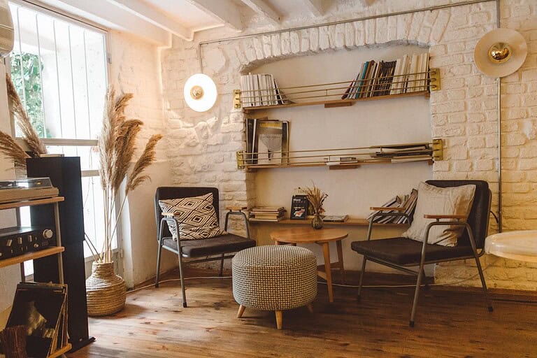 Cozy reading nook with white brick walls, two black chairs with patterned cushions, round knitted ottoman, brass bookshelves, wooden coffee table, pampas grass decoration, vinyl record player, warm natural lighting