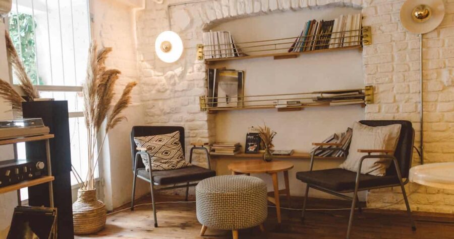 Cozy reading nook with white brick walls, two black chairs with patterned cushions, round knitted ottoman, brass bookshelves, wooden coffee table, pampas grass decoration, vinyl record player, warm natural lighting