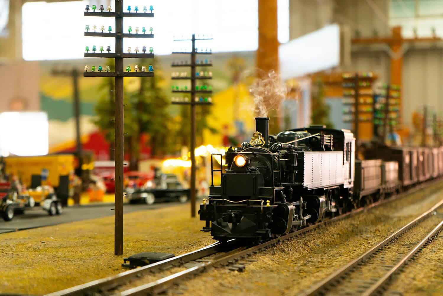 A model steam train moving along a track, with smoke rising from its chimney, surrounded by miniature trees, telephone poles with tiny figurines, and small cars, the scene depicting a detailed model railway setup