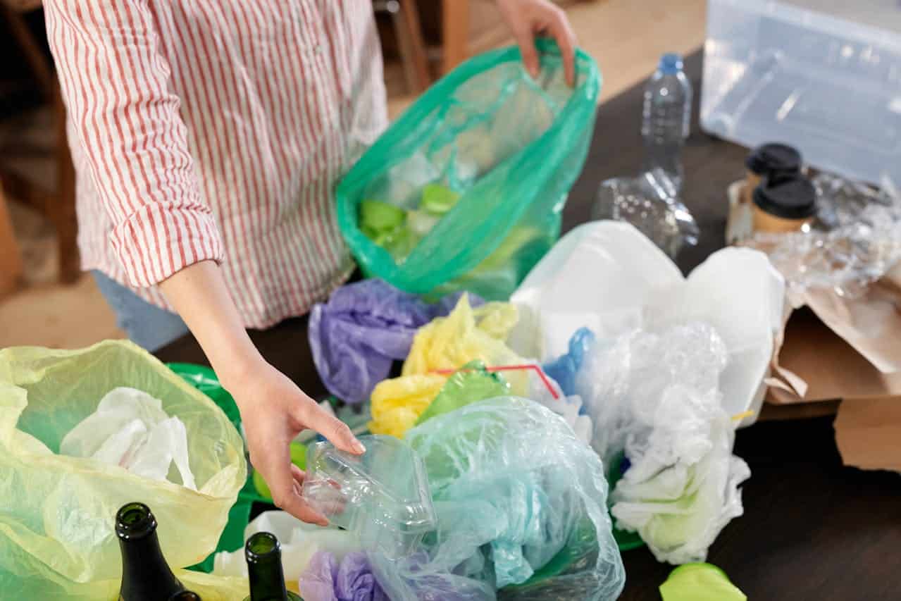 Person in red striped shirt sorting recyclable items, handling plastic packaging amid colorful bags. Table contains various plastic containers, bottles, and bags in green, yellow, purple, and white, showing waste separation or recycling activity