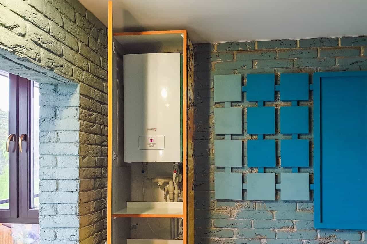Utility closet with water heater inside orange-framed cabinet, blue painted brick wall with decorative square tiles nearby