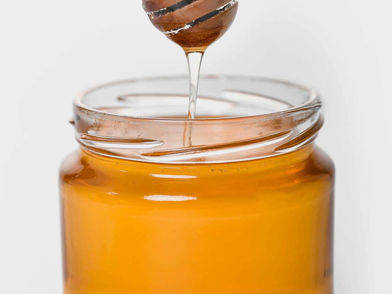 Honey dripping from a dipper into a clear glass jar, smooth golden texture, jar with a clean white background, natural lighting highlighting the honey's transparency, focus on the drips and the jar's opening, rich amber hue contrasting against the white backdrop