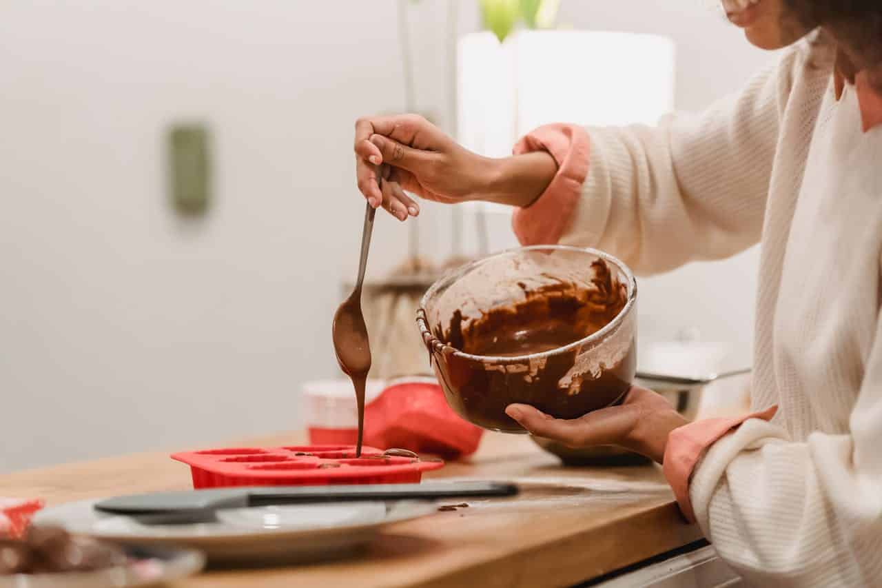 A person is pouring melted chocolate from a spoon into a red silicone mold while holding a bowl of chocolate, preparing to make homemade chocolates or desserts in a kitchen setting with soft lighting, the person is focused and wearing a casual sweater
