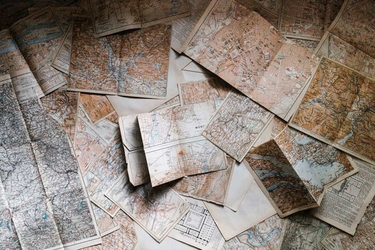 A collection of vintage maps spread out on a surface, featuring different regions and detailed topography, some maps partially overlapping, with faded colors and worn edges, creating a nostalgic and historical atmosphere