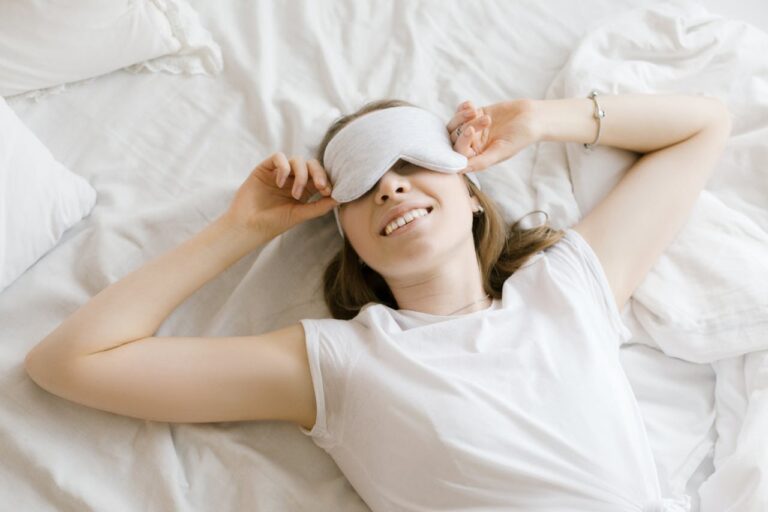 A smiling woman with a sleep mask on her eyes, lying on a white bed with soft sheets, she is removing the sleep mask from her face, her arm is resting comfortably, conveying a relaxed and peaceful mood