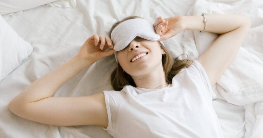 A smiling woman with a sleep mask on her eyes, lying on a white bed with soft sheets, she is removing the sleep mask from her face, her arm is resting comfortably, conveying a relaxed and peaceful mood