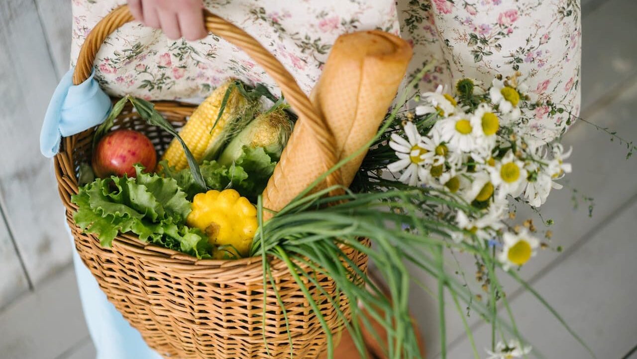 Person in floral dress holding wicker basket filled with fresh produce, bread, and daisies against light background