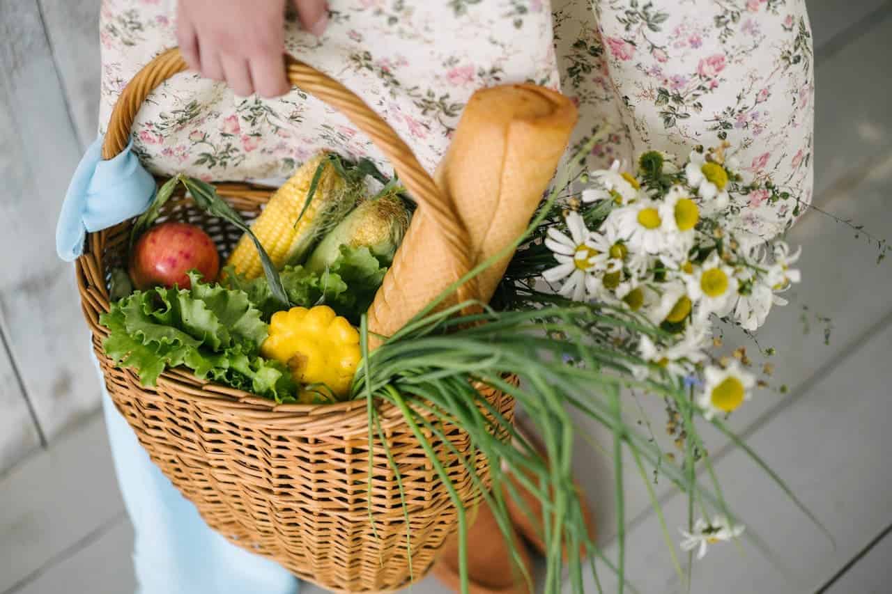 Person in floral dress holding wicker basket filled with fresh produce, bread, and daisies against light background
