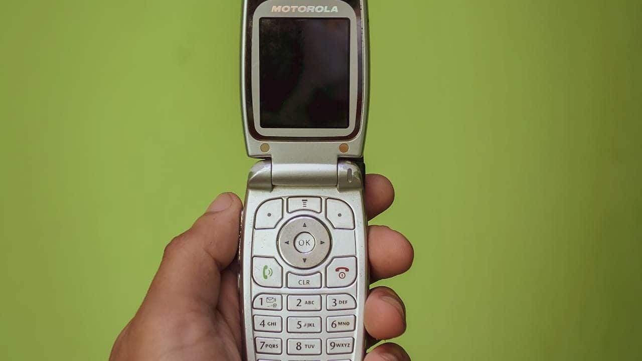 A hand holding an open Motorola flip phone, with visible buttons and a small screen, the phone's silver and white color contrasted against a bright green background, showcasing the design of a classic mobile phone