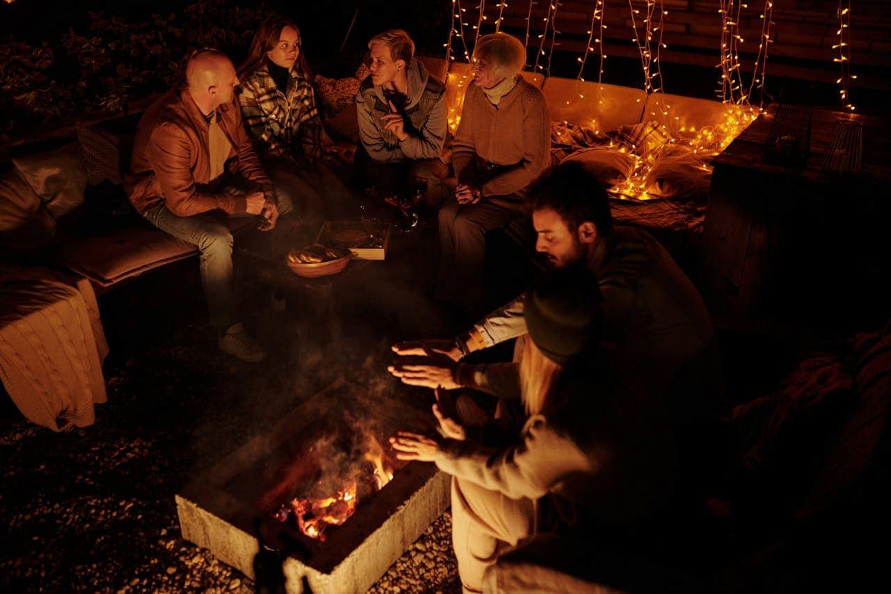 People gathered around outdoor fire pit at night, warming hands while seated on couches with string lights behind them