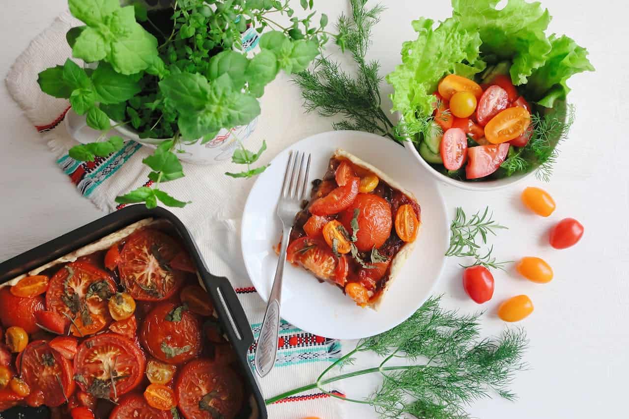Homemade tomato tart served with fresh salad, potted herbs, and scattered cherry tomatoes on embroidered cloth from overhead view