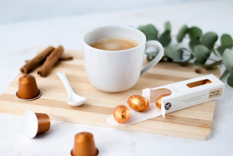 A cup of coffee placed on a wooden board, surrounded by cinnamon sticks, a small white spoon, chocolate-covered hazelnuts, and coffee capsules, the background is soft with a slight blur, the image conveys a cozy, relaxing moment for coffee lovers, with a touch of elegance from the chocolate packaging