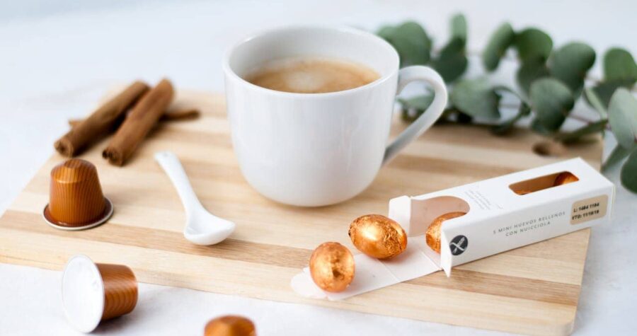 A cup of coffee placed on a wooden board, surrounded by cinnamon sticks, a small white spoon, chocolate-covered hazelnuts, and coffee capsules, the background is soft with a slight blur, the image conveys a cozy, relaxing moment for coffee lovers, with a touch of elegance from the chocolate packaging
