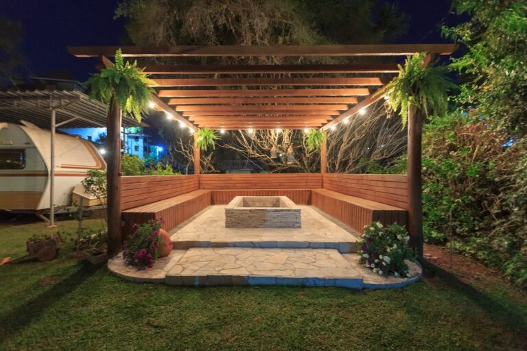 Wooden pergola with string lights covering stone patio featuring built-in bench seating and central fire pit at night