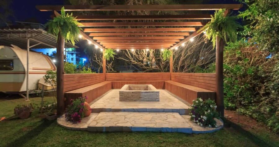 Wooden pergola with string lights covering stone patio featuring built-in bench seating and central fire pit at night