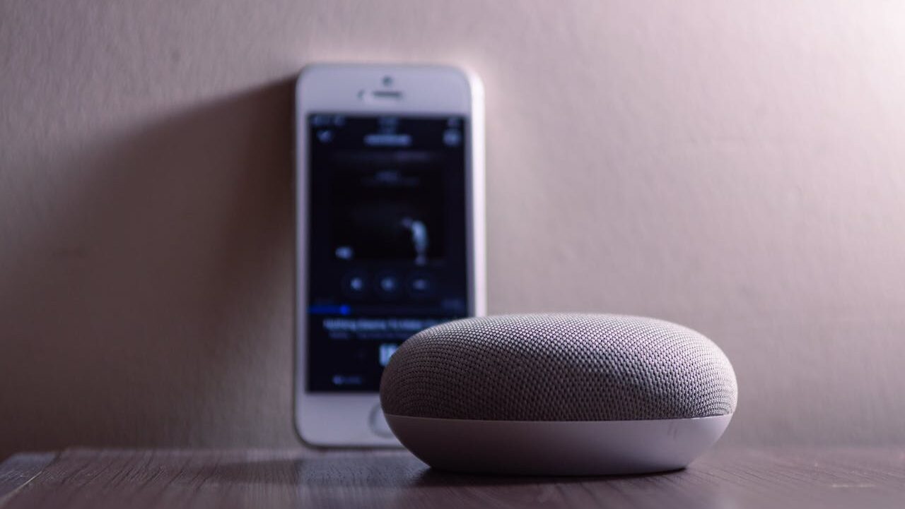 A smart speaker with a fabric exterior sits on a wooden table, an iPhone with a music streaming app is placed nearby, the phone screen displays song information, the image is lit softly, the focus is on the speaker, with a blurred background suggesting a modern, cozy atmosphere