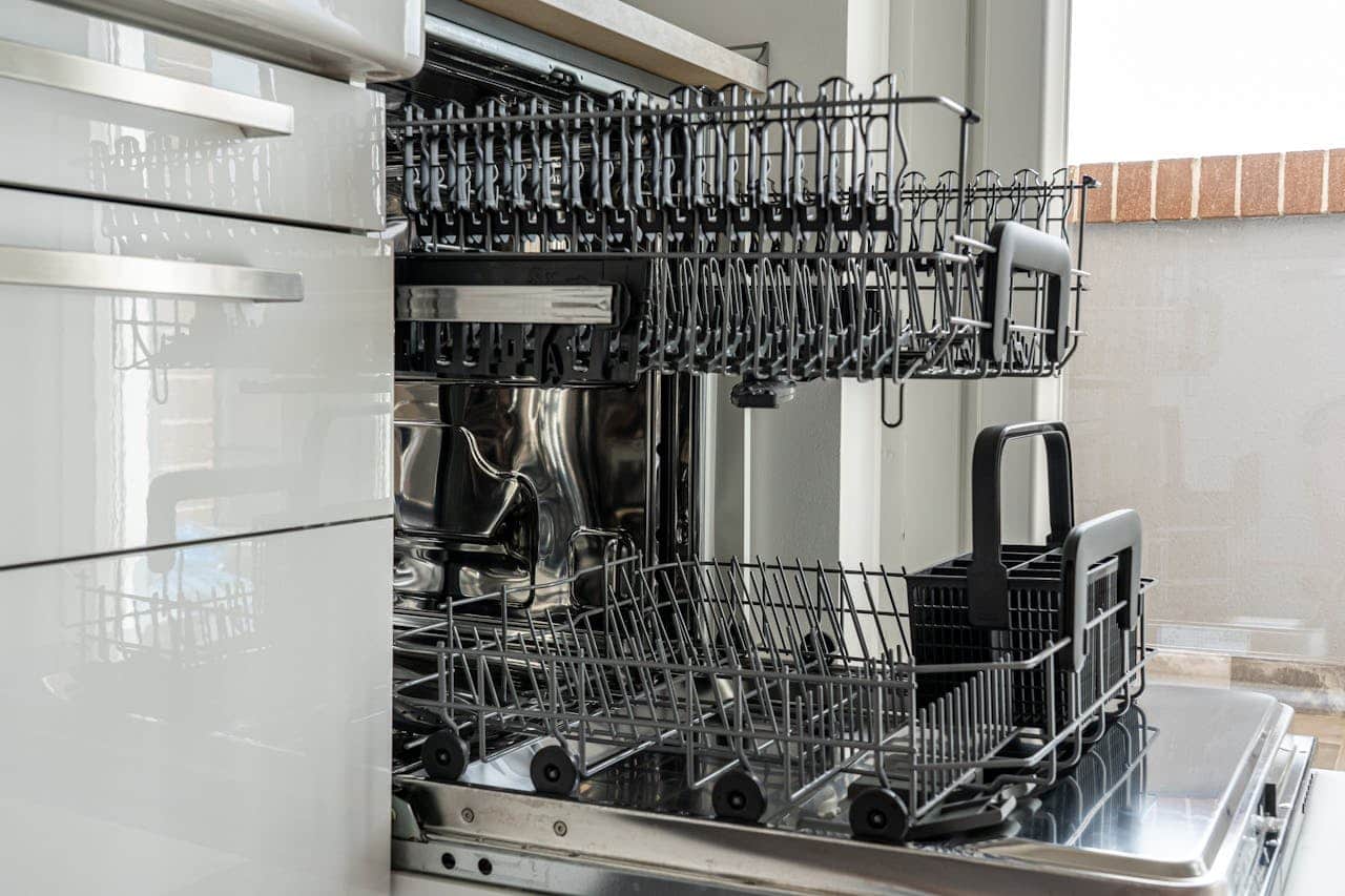 Open dishwasher showing empty interior with metal racks, utensil basket, and stainless steel interior in white kitchen cabinet