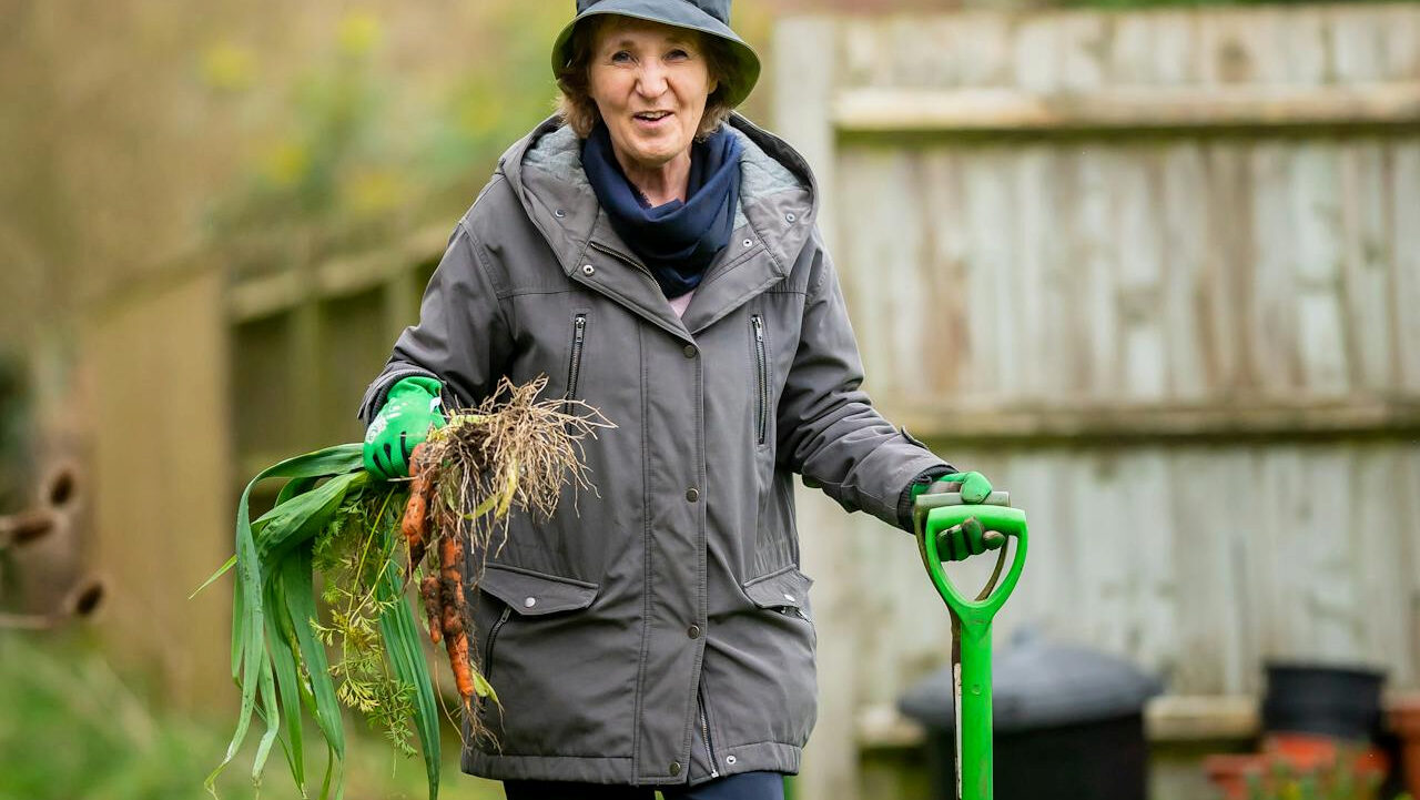 An elderly woman wearing a green coat and gloves, holding freshly harvested carrots in one hand and a green gardening tool in the other, smiling while standing in a garden with a wooden fence in the background, enjoying outdoor gardening