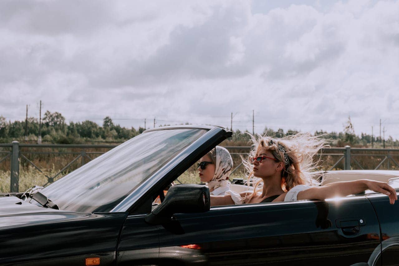 Two women are enjoying a drive in a convertible car, they are wearing sunglasses and scarves, the wind is blowing their hair, the sky is partly cloudy, and the background features a green landscape with a metal fence, the image conveys a relaxed, carefree vibe