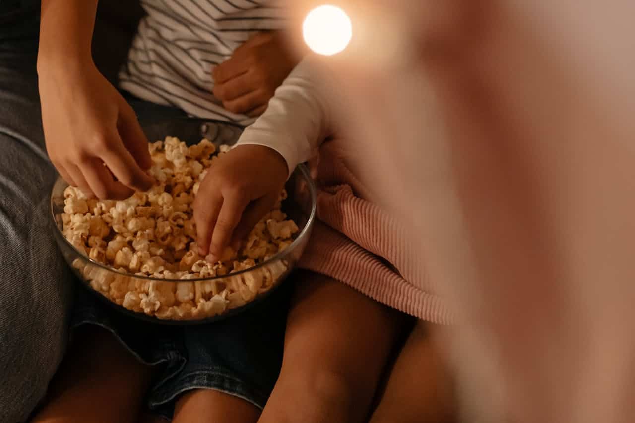 Two hands reaching into glass bowl of popcorn, people sitting together in dim lighting watching something