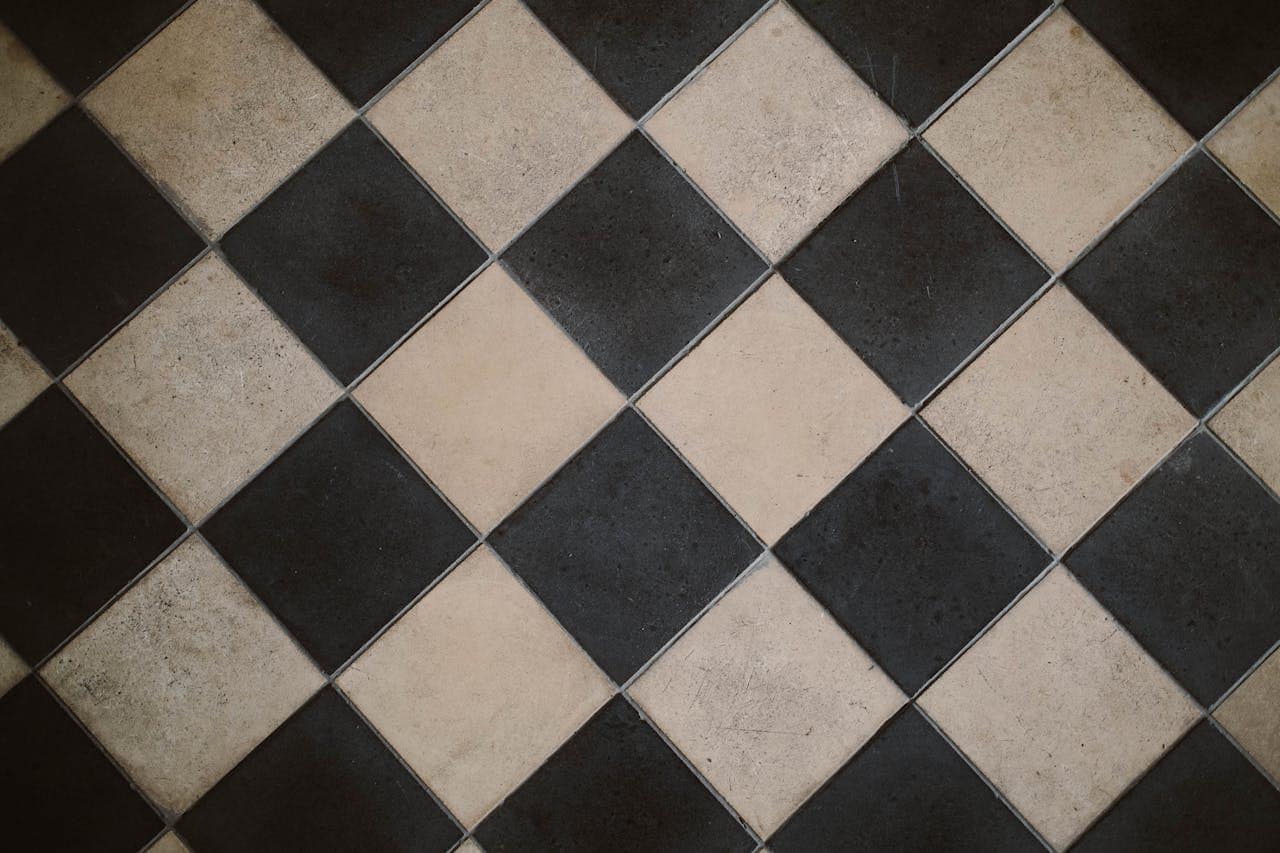 The image shows a floor with alternating black and beige tiles arranged in a diamond pattern, the tiles have visible wear and texture, creating a vintage aesthetic, geometric design across the floor