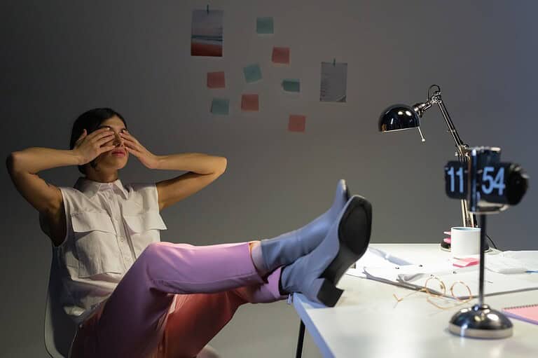 A person sitting at a desk, wearing pink pants and a white sleeveless shirt, with their hands covering their eyes, visibly tired or stressed, a clock showing 11:54, papers scattered on the desk, a desk lamp illuminating the scene, a few photos hanging on the wall, glasses placed on the desk