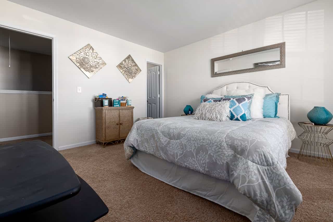 Bedroom with white tufted headboard, gray floral bedding, and teal accent pillows. Features rattan cabinet, metallic wall art, rectangular mirror, teal ceramic lamp, and wire side table on beige carpet