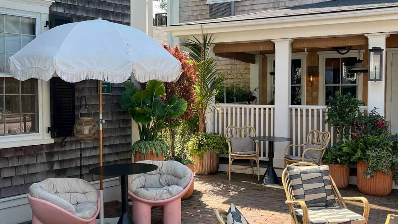 A cozy outdoor seating area with two pink cushioned chairs under a large white umbrella, a small black table between them, surrounded by potted plants, a wooden fence and balcony in the background, creating a peaceful and inviting space for relaxation or conversation