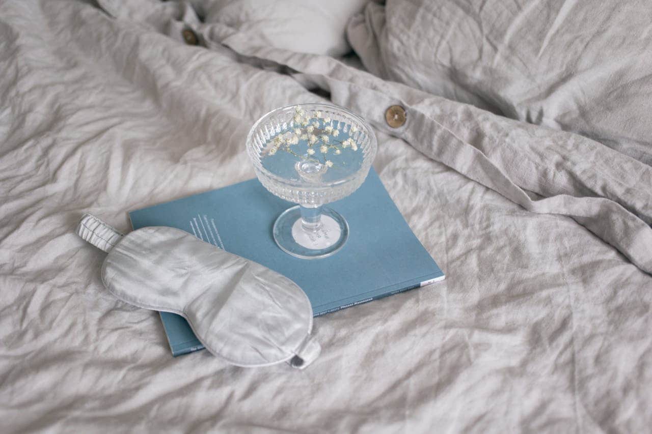 Crystal coupe glass containing blue liquid with small white flowers floating atop, placed on a light blue book with a white sleep mask, all resting on rumpled light gray cotton bedding