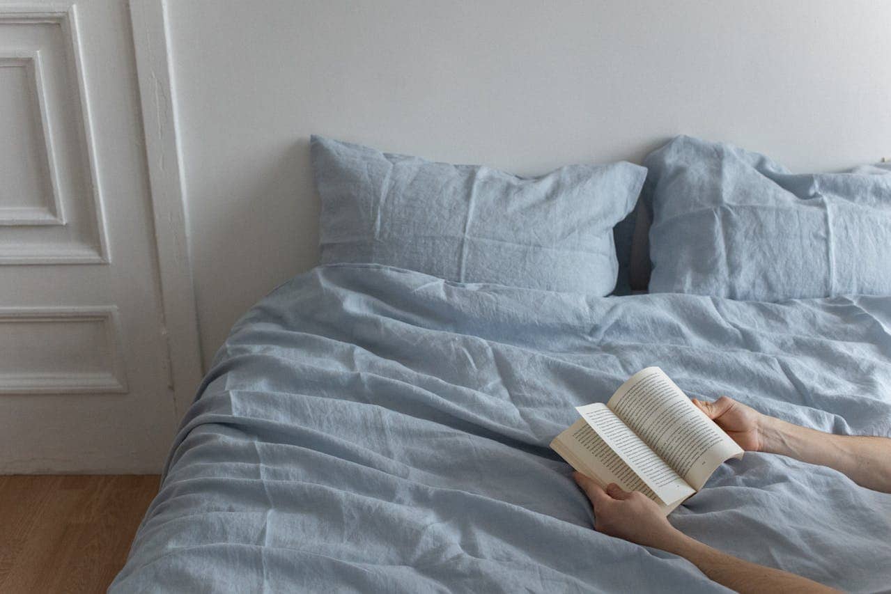 Person reading a book in bed with light blue linen bedding and matching pillows. White walls and paneled door visible. Hands holding open paperback against wrinkled duvet cover