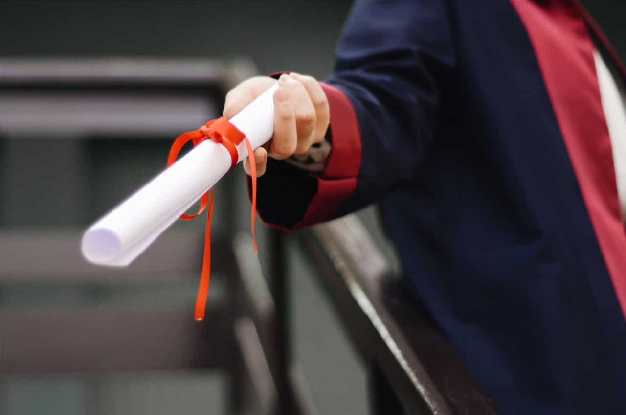 A person wearing a dark blue gown with a red collar, holding a rolled-up document tied with a red ribbon, the hand extended outward as if offering or presenting the document, symbolizing a graduation