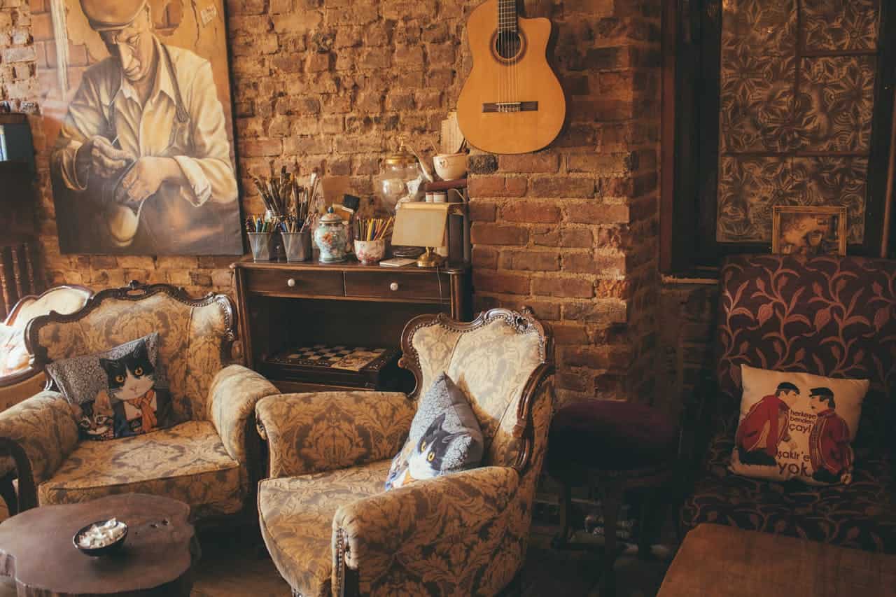 Cozy, vintage-inspired room with brick walls, two upholstered armchairs with cat-themed and couple pillows, a small wooden coffee table, a collection of colorful art supplies, a guitar hanging on the wall, and a warm, inviting atmosphere for relaxation and creativity
