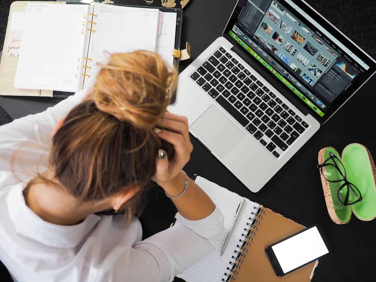 Person sitting at a desk with a laptop, holding their head in frustration, surrounded by notebooks, a phone, glasses, and an open planner, appears overwhelmed, focused on a task
