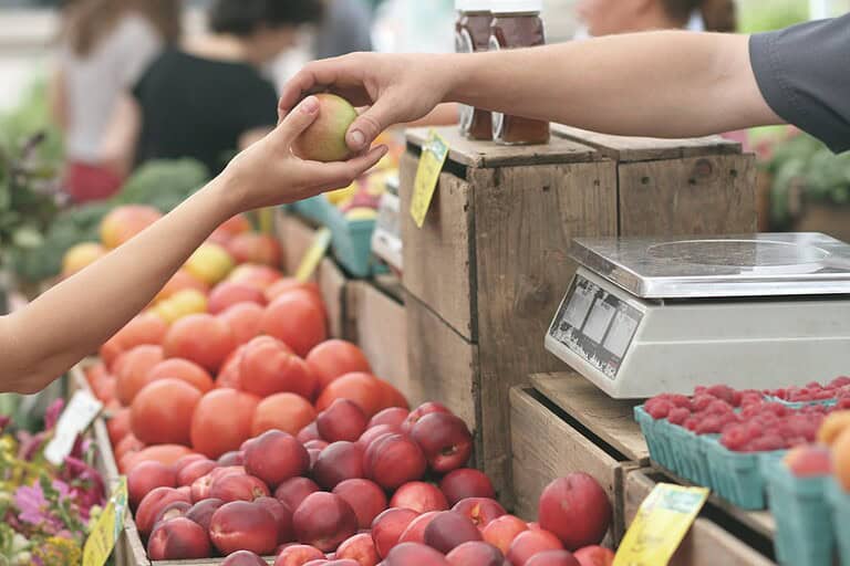 Two hands exchanging a green apple at a farmer's market, a variety of fresh fruits like peaches and tomatoes in the background, a scale is visible on the counter, the setting appears to be bright and outdoors, capturing a moment of purchase or transaction