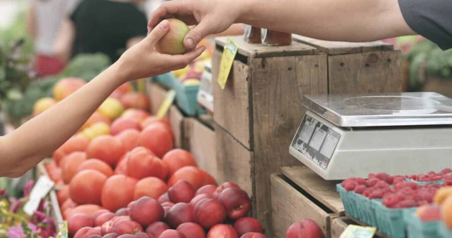 Two hands exchanging a green apple at a farmer's market, a variety of fresh fruits like peaches and tomatoes in the background, a scale is visible on the counter, the setting appears to be bright and outdoors, capturing a moment of purchase or transaction