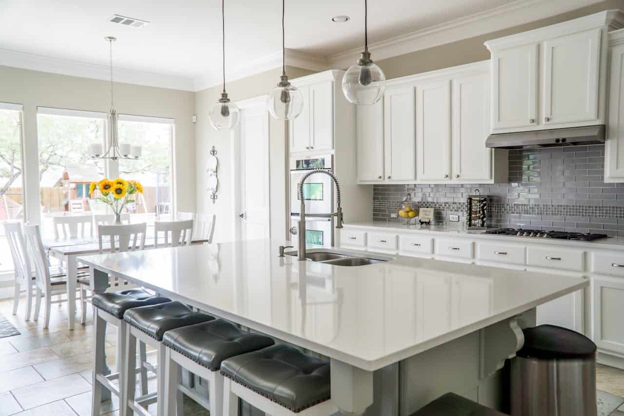 A modern, bright kitchen with white cabinetry, gray stone backsplash, stainless steel appliances, and a large island, a vase of sunflowers on the table, high stools at the island, natural light streaming through windows