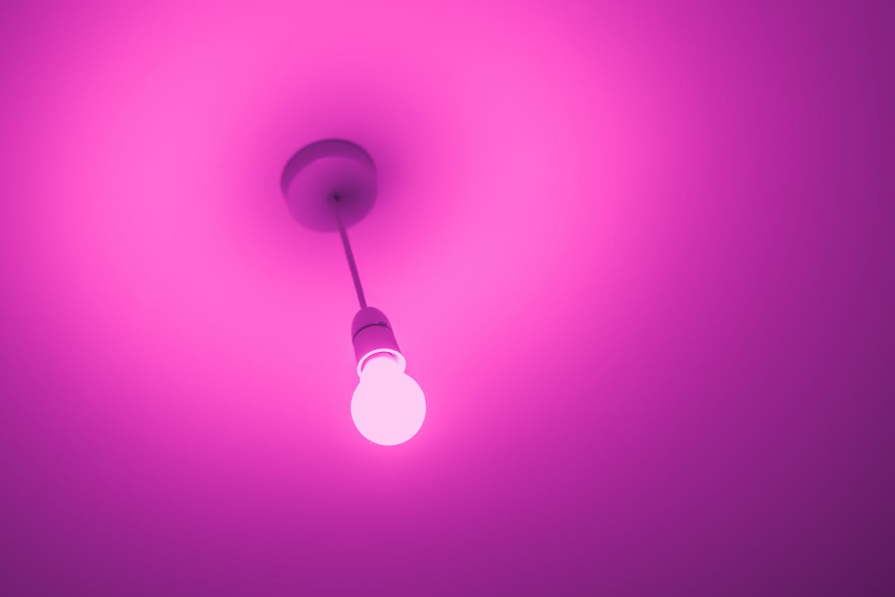 Pink light bulb glowing brightly from the ceiling, suspended by a thin cord, soft purple glow illuminating the space, light source in a minimalistic setting, modern and simple lighting fixture