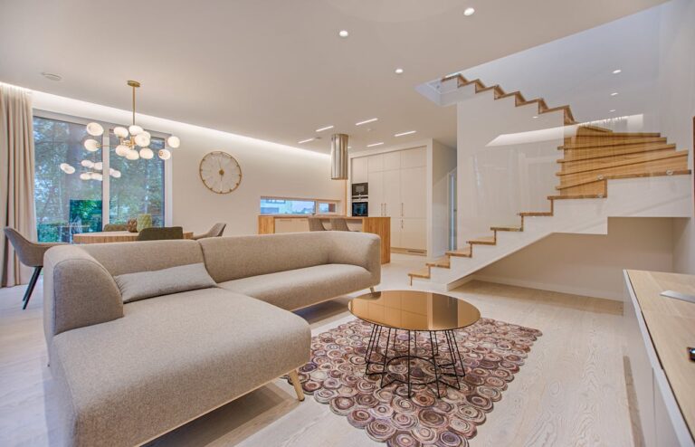 Modern open-concept living space with beige sectional sofa, wooden staircase with glass railings, spherical chandelier, and minimalist white kitchen