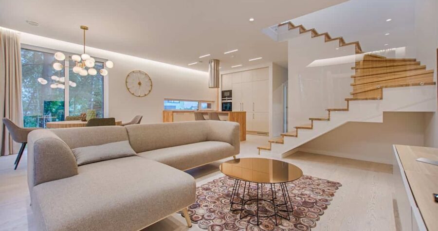 Modern open-concept living space with beige sectional sofa, wooden staircase with glass railings, spherical chandelier, and minimalist white kitchen