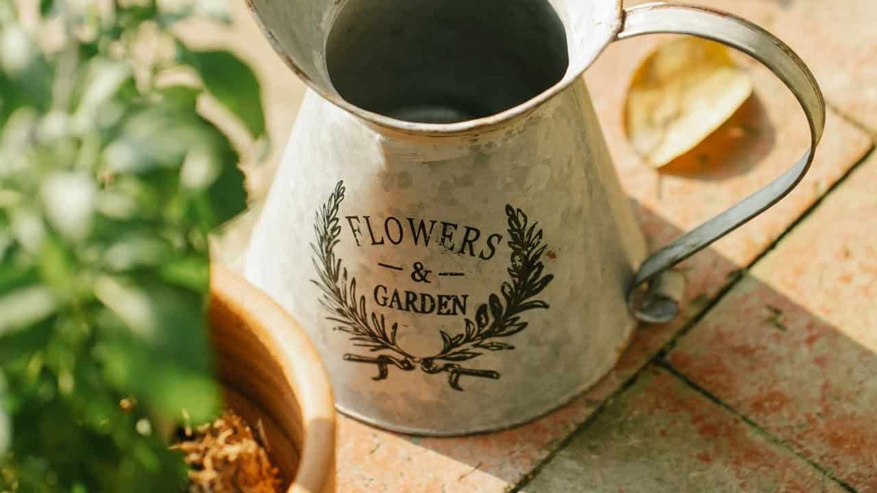 A metal watering can with the words "FLOWERS & GARDEN" printed on it, sitting next to a small flowerpot, the watering can is positioned at the forefront with a green plant visible in the background, the focus is on the intricate design, the setting appears serene and organized for gardening tasks