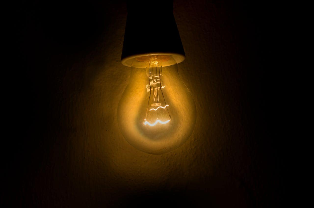 A glowing light bulb hanging from the ceiling, emitting warm yellow light, set against a dark background, soft illumination highlighting the filament inside, casting shadows around the bulb